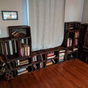 May include: A wall unit made of wooden crates, stacked and arranged to create a bookshelf. The crates are stained a dark brown color and are filled with books. Some of the crates are also used to store shoes.