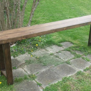 May include: A long, narrow, dark brown wooden console table featuring a smooth rectangular top surface and four sturdy, straight legs connected by horizontal support bars near the base, constructed from solid wood with a visible grain finish.