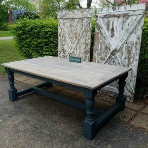 May include: A rectangular wooden table with a light gray stained top and dark charcoal blue painted legs and base. The table features four turned legs connected by a horizontal support beam. A small teal sign with the text Unique Primtiques sits on top.
