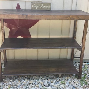 May include: A dark brown wooden shelf unit with three shelves. The shelf unit is standing on a gravel surface in front of a white wall. The sign above the shelf unit reads "Unique Printiques".