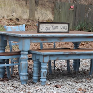 May include: A distressed blue dining set with a rectangular table, bench, and two chairs. The table has a brown top and turned legs. A sign on the table reads "Unique Primitives". The set is outdoors on a gravel surface.