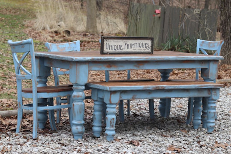May include: A distressed blue dining set with a rectangular table, bench, and two chairs. The table has a brown top and turned legs. A sign on the table reads "Unique Primitives". The set is outdoors on a gravel surface.