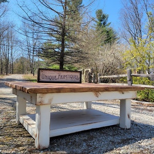 May include: White farmhouse style coffee table with a distressed finish. The table has a lower shelf and a wooden top with a sign that reads "Unique Primitives".