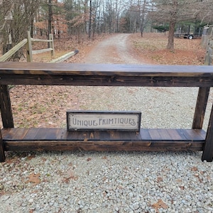 May include: A dark brown wooden console table with a lower shelf. The table has a distressed finish and a rustic look. A small sign with the text "Unique Primitives" is sitting on the shelf.