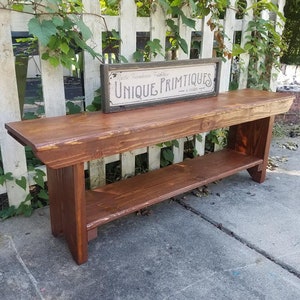 May include: A rustic wooden bench with a shelf underneath. The bench is stained a dark brown color and has a distressed look. The bench is sitting in front of a white picket fence with green vines growing on it. A sign that reads "Unique Primitives" is sitting on top of the bench.