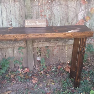May include: A dark brown wooden console table with a rustic finish. The table has a rectangular top and two legs on each side. The table is sitting in front of a wooden fence with vines growing on it. A small wooden sign with the text "Unique Finds" is sitting on the table.
