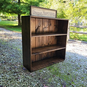 May include: A dark brown wooden shelf unit with three shelves. The shelf unit is sitting on a gravel surface. A sign above the shelf unit reads "Unique Primitives".
