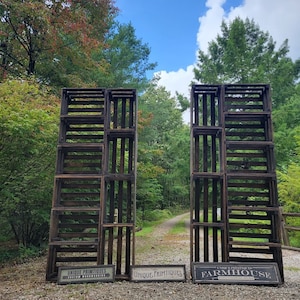 May include: Two wooden crate displays with signs that read "Unique Primitives Custom Handcrafted" and "Unique Primitives Farmhouse". The crates are stacked in a rustic setting with a dirt path and trees in the background.