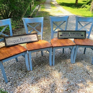 May include: Four light blue wooden chairs with brown seats. The chairs have an X-shaped design on the back. Two signs with the text "UNIQUE PRIMTIQUES" are placed on the seats. The chairs are on a gravel surface.