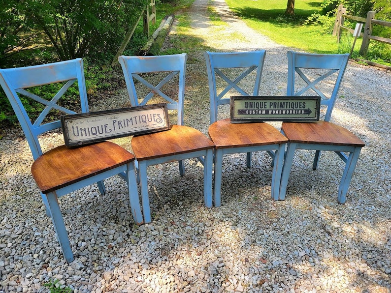 May include: Four light blue wooden chairs with brown seats. The chairs have an X-shaped design on the back. Two signs with the text "UNIQUE PRIMTIQUES" are placed on the seats. The chairs are on a gravel surface.