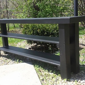 May include: Black wooden console table with two shelves. The table is made of solid wood and has a rustic finish. It is perfect for displaying decor or storing items.