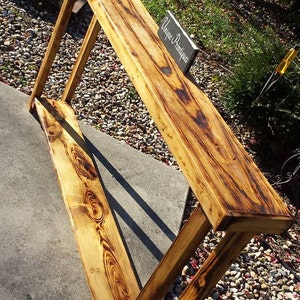 May include: A rustic wooden console table with a distressed, burnt wood finish. The table has a single shelf and is made of solid wood. The table is sitting on a gravel surface with a sign that reads "Unique Antiques".