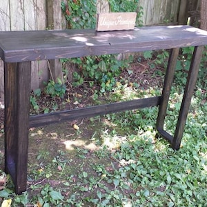 May include: A black wooden console table with a simple, modern design. The table has a rectangular top and two vertical supports with a horizontal bar connecting them. The table is sitting on a grassy surface.