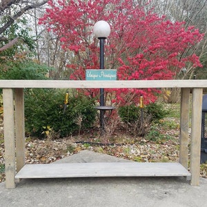 May include: A rustic wooden console table with a gray weathered finish. The table has a single shelf and is made of reclaimed wood. The table is in front of a red bush and a black metal fire pit.