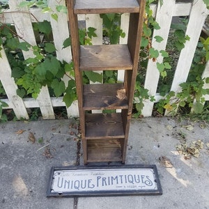 May include: A wooden shelf unit with four shelves, stained a dark brown color. The shelf unit is standing on a gray concrete surface. A sign that reads "Rustic Farmhouse Furniture Unique Primitives & Decor" is lying on the ground in front of the shelf unit.