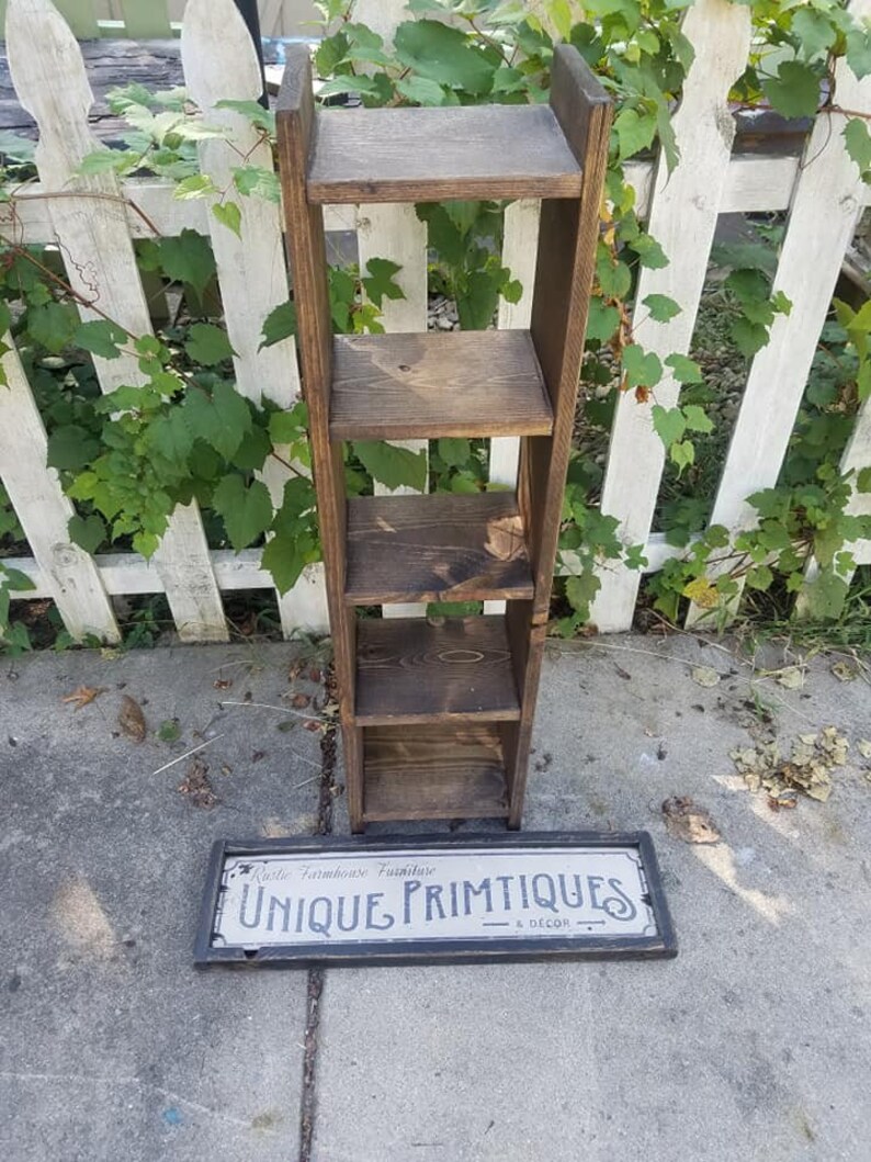 May include: A wooden shelf unit with four shelves, stained a dark brown color. The shelf unit is standing on a gray concrete surface. A sign that reads "Rustic Farmhouse Furniture Unique Primitives & Decor" is lying on the ground in front of the shelf unit.