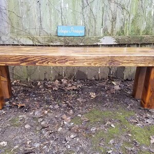 May include: A rustic wooden bench with a rich, warm brown stain. The bench features a thick, rectangular seat supported by four sturdy, rectangular legs. The wood grain is visible, adding to the natural aesthetic. A small sign in the background reads "Unique Primatiques".