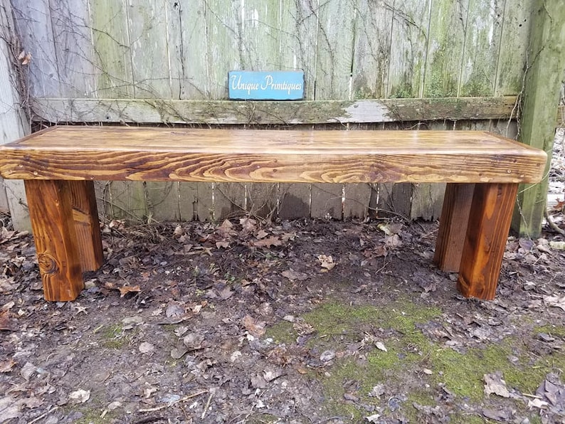 May include: A rustic wooden bench with a rich, warm brown stain. The bench features a thick, rectangular seat supported by four sturdy, rectangular legs. The wood grain is visible, adding to the natural aesthetic. A small sign in the background reads "Unique Primatiques".