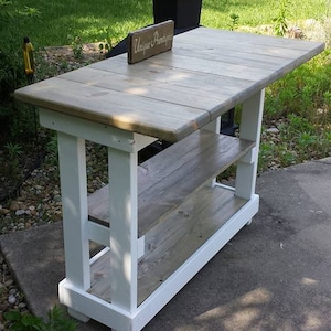 May include: White painted wooden table with two shelves and a gray wood top. The table has a sign that says "Unique Printshop" on it.