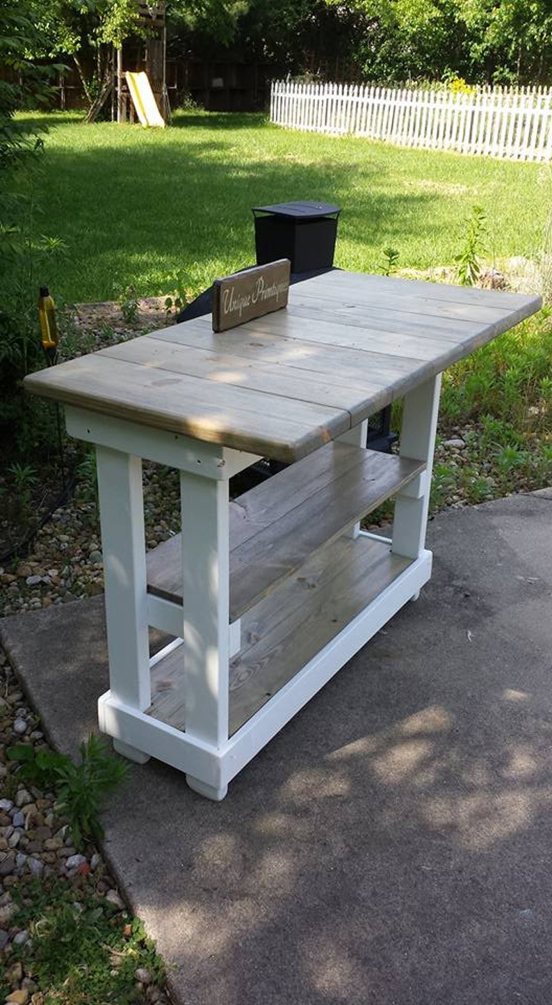 May include: White painted wooden table with two shelves and a gray wood top. The table has a sign that says "Unique Printshop" on it.