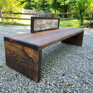 May include: A rustic wooden bench with a dark brown stain. The bench has a wide, flat top and a slightly curved underside. It is sitting on a gravel driveway with a vintage sign that reads "Unique Primitives" in the background.