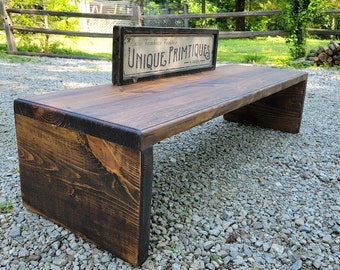 May include: A rustic wooden bench with a dark brown stain. The bench has a wide, flat top and a slightly curved underside. It is sitting on a gravel driveway with a vintage sign that reads "Unique Primitives" in the background.