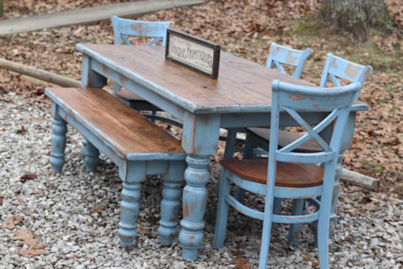 May include: A rustic dining set with a distressed blue finish. The set includes a rectangular table, a bench, and three chairs. The table has a brown wooden top and a sign that reads "UNIQUE PRIMTIQUES".
