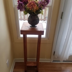 May include: A wooden plant stand with a square top and two vertical supports. A vase with a bouquet of purple, pink, and yellow flowers sits on the top of the stand. The stand is in front of a window with a view of trees and a body of water. A sign with the text "Unique Primitives" is on the floor in front of the stand.