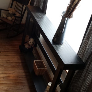 May include: A black wooden console table with two shelves. The table has a distressed finish and is decorated with a vase of dried flowers, a stuffed animal, and a lantern.