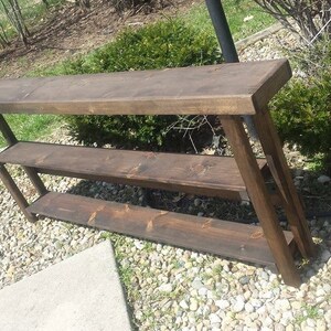 May include: A dark brown wooden console table with three shelves. The table is made of reclaimed wood and has a rustic finish.
