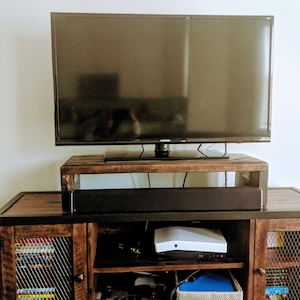 May include: A dark wood entertainment center with a black metal frame. A flat screen TV sits on a wooden shelf above a black sound bar. A white streaming device is on the lower shelf.