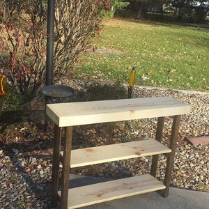 May include: A wooden console table with two shelves. The tabletop and shelves are light-colored wood, supported by dark brown legs. The table is outdoors, set on a concrete surface with a backdrop of greenery and a lamp post.