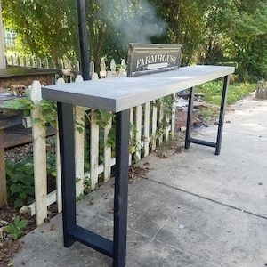 May include: A long, gray, rectangular table with black metal legs. The table is sitting on a concrete surface. A sign that says "Unusually Painted Farmhouse" is sitting on the table.