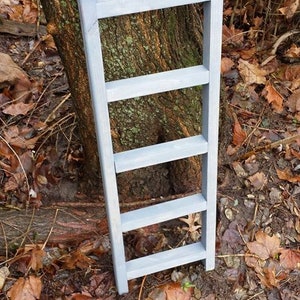 May include: A gray wooden ladder with five rungs, standing on a bed of brown leaves.