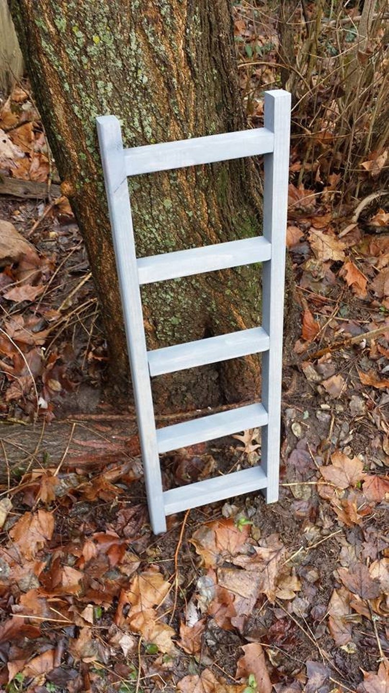 May include: A gray wooden ladder with five rungs, standing on a bed of brown leaves.