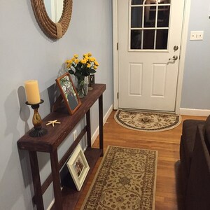 May include: A dark brown wooden console table with a shelf, featuring a framed photo, a candle holder with a lit candle, a starfish, and a vase of yellow flowers. The table is placed in a hallway with a light blue wall, a white door, and a patterned rug.