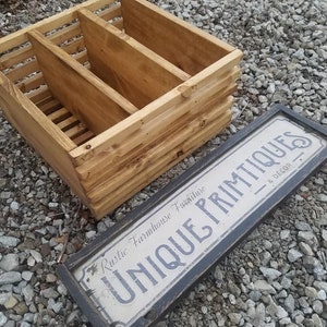 May include: A wooden crate with three compartments, stained in a light brown color. The crate is sitting on a bed of small gray rocks. A black framed sign with white lettering reads "Unique Primitives - Rustic Farmhouse Furniture & Decor".
