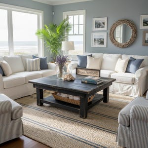 May include: A square black wooden coffee table with a lower shelf holding a woven basket of white towels. The table surface features a vase of lavender, a decorative sign reading Unique Primtiques, and stacked books in a living room setting.