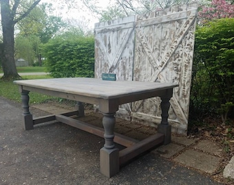May include: A rectangular wooden dining table with a light gray tabletop and dark gray turned legs. The table features a sturdy base with horizontal support beams connecting the four legs. A small teal sign is placed on the center of the table.