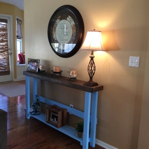 May include: A light blue console table with a wooden top and a shelf. The table has a lamp with a white shade and a framed picture on top. The wall behind the table is a light beige color.