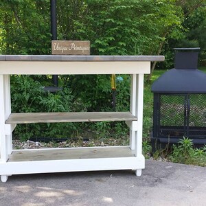 May include: White wooden table with two shelves and a gray top. The table has a sign that says "Unique  Primitives" on it. A black metal fire pit is to the right of the table.