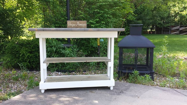 May include: White wooden table with two shelves and a gray top. The table has a sign that says "Unique  Primitives" on it. A black metal fire pit is to the right of the table.