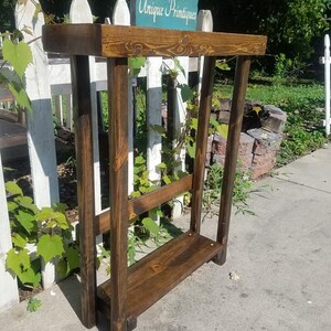 May include: A dark brown wooden console table with a single shelf. The table is made of rustic wood and has a distressed finish. The table is standing on a concrete surface. A sign that reads "Unique Primtiques" is visible in the background.