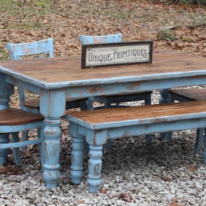 May include: A rustic dining set with a distressed blue finish. The set includes a rectangular table, a bench, and chairs. The table has a wooden top and the sign on the table reads "UNIQUE PRIMTIQUES". The chairs have an X-back design.