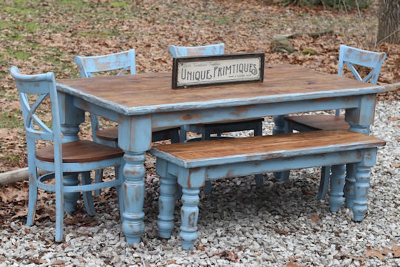 May include: A rustic dining set with a distressed blue finish. The set includes a rectangular table, a bench, and chairs. The table has a wooden top and the sign on the table reads "UNIQUE PRIMTIQUES". The chairs have an X-back design.