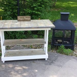 May include: A white wooden table with two shelves and a gray wooden top. The table has a sign that says "Unique Primitive" on it. A black metal fire pit is in the background.