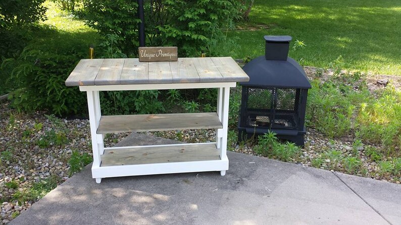 May include: A white wooden table with two shelves and a gray wooden top. The table has a sign that says "Unique Primitive" on it. A black metal fire pit is in the background.