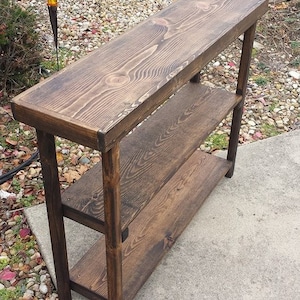 May include: A dark brown wooden console table with two shelves. The table has a rustic, distressed finish and is made of solid wood.