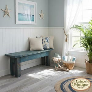 May include: A dark teal wooden bench with a rectangular seat and four sturdy legs, positioned against a white paneled wall. Two square throw pillows, one solid beige and one with a blue coral pattern, rest on the bench seat.
