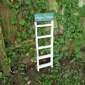 May include: White wooden ladder shelf with five shelves, standing in front of a green ivy covered wooden fence. A teal blue sign with the text "Unique Primitives" is attached to the top of the shelf.